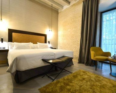 Habitación contemporánea en el Hotel Legado Alcázar de Sevilla, que cuenta con una cama grande, una silla mostaza, una mesa y una espaciosa ventana con cortinas oscuras, bellamente iluminada por lámparas de pared. - ForTwo - Planes para parejas