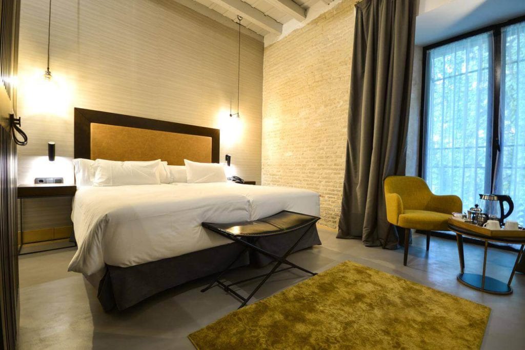 Habitación contemporánea en el Hotel Legado Alcázar de Sevilla, que cuenta con una cama grande, una silla mostaza, una mesa y una espaciosa ventana con cortinas oscuras, bellamente iluminada por lámparas de pared. - ForTwo - Planes para parejas