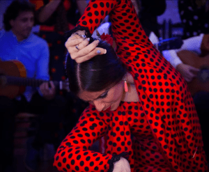 En medio de las animadas Cuevas del Sacromonte, el "Espectáculo Flamenco en las Cuevas del Sacromonte con Los Tarantos" muestra a una intensa bailaora de flamenco con un vestido de lunares rojos y negros, rodeada de músicos. - ForTwo - Planes para parejas