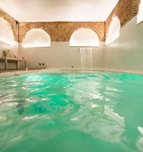 Relájese en el spa del Hotel Hospes Puerta de Alcalá, Madrid, con aguas turquesas bajo un techo abovedado de ladrillo. Una cascada ofrece sonidos relajantes y la luz de las velas enriquece la atmósfera serena. - ForTwo - Planes para parejas