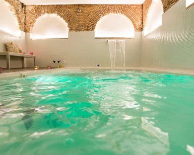 Relájese en el spa del Hotel Hospes Puerta de Alcalá, Madrid, con aguas turquesas bajo un techo abovedado de ladrillo. Una cascada ofrece sonidos relajantes y la luz de las velas enriquece la atmósfera serena. - ForTwo - Planes para parejas