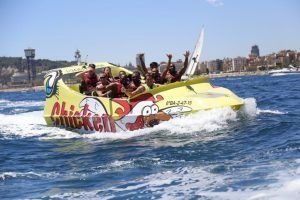 Un grupo disfruta de las vibrantes olas mientras corren por las aguas cercanas al impresionante paisaje urbano de Barcelona en "Surca las olas a toda velocidad en Chicken Boat en Barcelona". - ForTwo - Planes para parejas