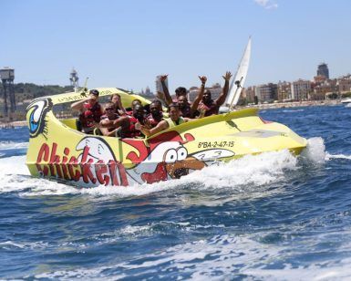 Un grupo disfruta de las vibrantes olas mientras corren por las aguas cercanas al impresionante paisaje urbano de Barcelona en "Surca las olas a toda velocidad en Chicken Boat en Barcelona". - ForTwo - Planes para parejas