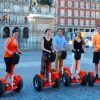 En una plaza bulliciosa, cinco personas se deslizan sin esfuerzo en Segways rojos bajo la belleza escénica del Madrid histórico, con sus grandes edificios y estatuas. - ForTwo - Planes para parejas