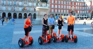 En una plaza bulliciosa, cinco personas se deslizan sin esfuerzo en Segways rojos bajo la belleza escénica del Madrid histórico, con sus grandes edificios y estatuas. - ForTwo - Planes para parejas