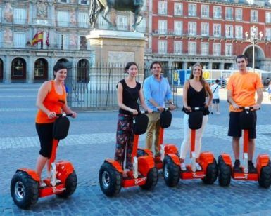 En una plaza bulliciosa, cinco personas se deslizan sin esfuerzo en Segways rojos bajo la belleza escénica del Madrid histórico, con sus grandes edificios y estatuas. - ForTwo - Planes para parejas