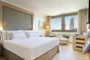 Disfrute de una moderna habitación del Hotel Meliá Princesa (Madrid) con una cama grande, una pequeña mesa redonda con dos sillas y una ventana que muestra impresionantes vistas del dinámico paisaje urbano de Madrid. - ForTwo - Planes para parejas