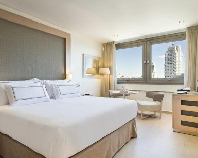 Disfrute de una moderna habitación del Hotel Meliá Princesa (Madrid) con una cama grande, una pequeña mesa redonda con dos sillas y una ventana que muestra impresionantes vistas del dinámico paisaje urbano de Madrid. - ForTwo - Planes para parejas