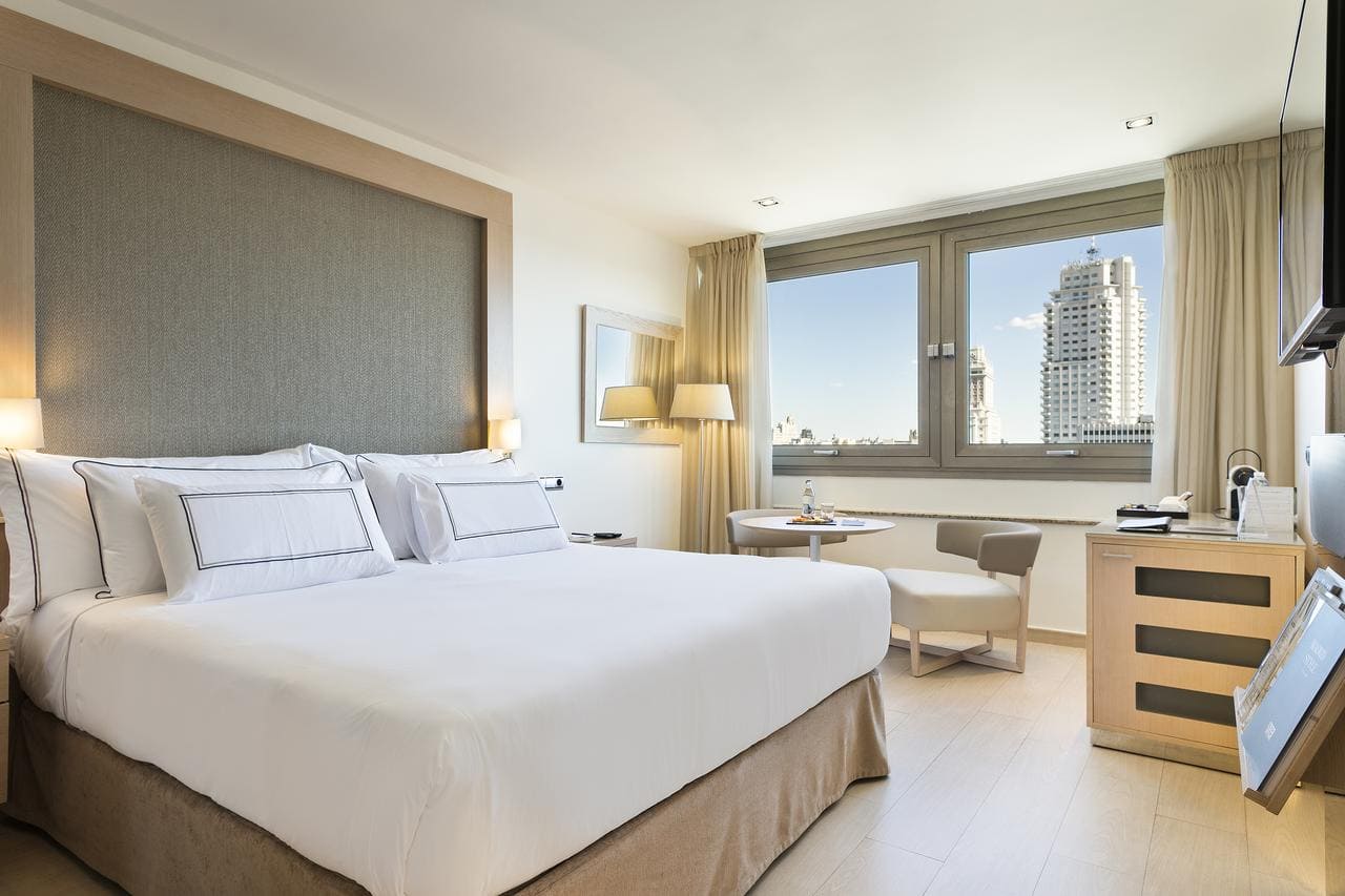 Disfrute de una moderna habitación del Hotel Meliá Princesa (Madrid) con una cama grande, una pequeña mesa redonda con dos sillas y una ventana que muestra impresionantes vistas del dinámico paisaje urbano de Madrid. - ForTwo - Planes para parejas