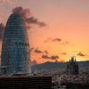 Un paisaje urbano que presenta la singular torre de cristal Mirador Torre Glorias junto a una catedral al atardecer, bajo un cielo adornado con nubes rosas y naranjas. - ForTwo - Planes para parejas