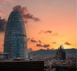 Un paisaje urbano que presenta la singular torre de cristal Mirador Torre Glorias junto a una catedral al atardecer, bajo un cielo adornado con nubes rosas y naranjas. - ForTwo - Planes para parejas