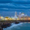 El paisaje urbano costero de Cádiz, con una catedral histórica bajo un cielo nublado espectacular, brilla al anochecer con edificios iluminados que susurran sobre la "Visita guiada de los misterios y leyendas de Cádiz". - ForTwo - Planes para parejas