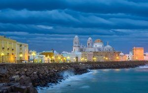 El paisaje urbano costero de Cádiz, con una catedral histórica bajo un cielo nublado espectacular, brilla al anochecer con edificios iluminados que susurran sobre la "Visita guiada de los misterios y leyendas de Cádiz". - ForTwo - Planes para parejas