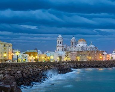 El paisaje urbano costero de Cádiz, con una catedral histórica bajo un cielo nublado espectacular, brilla al anochecer con edificios iluminados que susurran sobre la "Visita guiada de los misterios y leyendas de Cádiz". - ForTwo - Planes para parejas