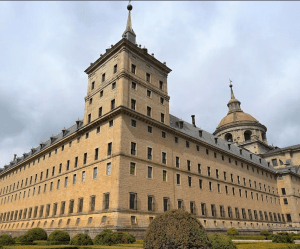 Explora un histórico edificio de piedra con ventanas arqueadas, techo abovedado y arbustos bien cuidados bajo un cielo nublado. Complementa tu visita con "Entradas al Monasterio del Escorial: Visita guiada" para apreciar plenamente su impresionante arquitectura. - ForTwo - Planes para parejas