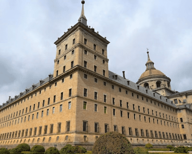 Explora un histórico edificio de piedra con ventanas arqueadas, techo abovedado y arbustos bien cuidados bajo un cielo nublado. Complementa tu visita con "Entradas al Monasterio del Escorial: Visita guiada" para apreciar plenamente su impresionante arquitectura. - ForTwo - Planes para parejas