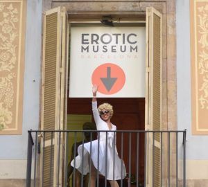Una persona con vestido blanco y peluca saluda desde un balcón bajo el cartel de Entradas Museo Erótico de Barcelona, capturando el espíritu lúdico de la ciudad. - ForTwo - Planes para parejas