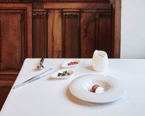 La elegante mesa del Restaurante Novodabo de Zaragoza muestra su exquisito Menú degustación con un plato blanco, una jarra pequeña, cubiertos y guarniciones sobre un mantel impecable entre cálidas paredes de madera. - ForTwo - Planes para parejas