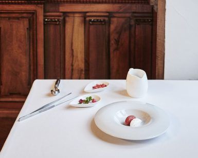 La elegante mesa del Restaurante Novodabo de Zaragoza muestra su exquisito Menú degustación con un plato blanco, una jarra pequeña, cubiertos y guarniciones sobre un mantel impecable entre cálidas paredes de madera. - ForTwo - Planes para parejas