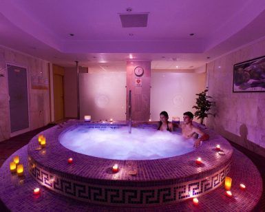Una pareja se relaja en un jacuzzi circular rodeado de velas encendidas en una sala de spa tenuemente iluminada, evocando el ambiente sereno de la experiencia del "Circuito termal de spa con masaje, té, chocolate y fruta" en Tenerife. - ForTwo - Planes para parejas