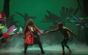 Actores con trajes coloridos dramatizan una escena en el escenario del Teatro Maravillas, rodeados de plantas vibrantes, dando vida a la magia de "Peter Pan, el musical". - ForTwo - Planes para parejas