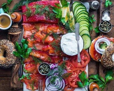 Fotografía cenital de un brunch en el restaurante Brunch en Picnic de Barcelona, con bagels, salmón ahumado, queso crema, alcaparras, tomates, pepinos, café y hierbas en una mesa de madera. - ForTwo - Planes para parejas
