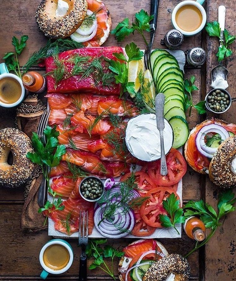Fotografía cenital de un brunch en el restaurante Brunch en Picnic de Barcelona, con bagels, salmón ahumado, queso crema, alcaparras, tomates, pepinos, café y hierbas en una mesa de madera. - ForTwo - Planes para parejas