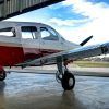 En un hangar, una pequeña avioneta roja y blanca espera, con las puntas de sus alas brillando a la luz del sol. Perfectamente preparada para el "Vuelo en avioneta a Barcelona", imagina el emocionante viaje por los vibrantes paisajes de Barcelona. - ForTwo - Planes para parejas