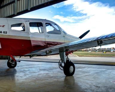 En un hangar, una pequeña avioneta roja y blanca espera, con las puntas de sus alas brillando a la luz del sol. Perfectamente preparada para el "Vuelo en avioneta a Barcelona", imagina el emocionante viaje por los vibrantes paisajes de Barcelona. - ForTwo - Planes para parejas