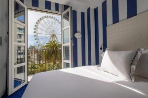El Hotel Room Mate Valeria Málaga ofrece una encantadora habitación con paredes de rayas azules y puertas de balcón abiertas, que ofrecen una vista impresionante de una noria y palmeras. - ForTwo - Planes para parejas