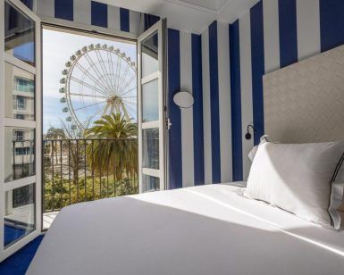 El Hotel Room Mate Valeria Málaga ofrece una encantadora habitación con paredes de rayas azules y puertas de balcón abiertas, que ofrecen una vista impresionante de una noria y palmeras. - ForTwo - Planes para parejas