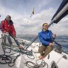 Una pareja con chaqueta y gafas de sol navega en un elegante velero de carreras francés bajo el sol de Barcelona, con nubes esponjosas como telón de fondo. - ForTwo - Planes para parejas