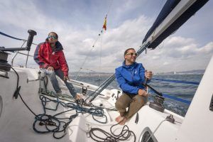 Una pareja con chaqueta y gafas de sol navega en un elegante velero de carreras francés bajo el sol de Barcelona, con nubes esponjosas como telón de fondo. - ForTwo - Planes para parejas