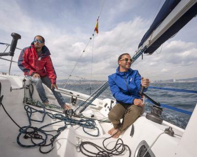Una pareja con chaqueta y gafas de sol navega en un elegante velero de carreras francés bajo el sol de Barcelona, con nubes esponjosas como telón de fondo. - ForTwo - Planes para parejas