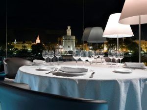 Cene elegantemente en el Restaurante Abades Triana con una mesa preparada para seis personas, disfrutando de una vista nocturna del paisaje urbano iluminado y la histórica Torre de Triana. - ForTwo - Planes para parejas