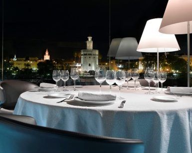 Cene elegantemente en el Restaurante Abades Triana con una mesa preparada para seis personas, disfrutando de una vista nocturna del paisaje urbano iluminado y la histórica Torre de Triana. - ForTwo - Planes para parejas