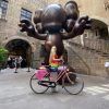 Una persona en bicicleta frente a una gran escultura marrón tipo caricatura afuera de un edificio de piedra se mezcla con el vibrante arte callejero de Barcelona en un inolvidable Barcelona Street Art Tour. - ForTwo - Planes para parejas