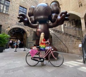 Una persona en bicicleta frente a una gran escultura marrón tipo caricatura afuera de un edificio de piedra se mezcla con el vibrante arte callejero de Barcelona en un inolvidable Barcelona Street Art Tour. - ForTwo - Planes para parejas