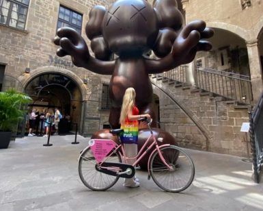 Una persona en bicicleta frente a una gran escultura marrón tipo caricatura afuera de un edificio de piedra se mezcla con el vibrante arte callejero de Barcelona en un inolvidable Barcelona Street Art Tour. - ForTwo - Planes para parejas