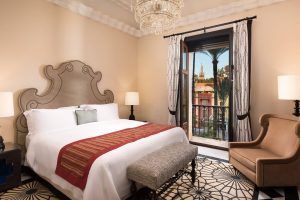 Descubra la máxima elegancia en la Suite Deluxe del Hotel Alfonso XIII de Sevilla. Disfrute de una espaciosa habitación con una cama grande, lámpara de araña y sillón, mientras admira las impresionantes vistas a una histórica torre enmarcada por frondosas palmeras desde la ventana. - ForTwo - Planes para parejas