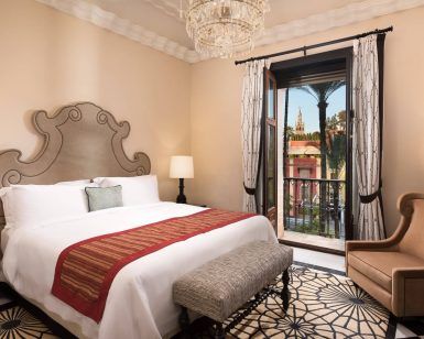 Descubra la máxima elegancia en la Suite Deluxe del Hotel Alfonso XIII de Sevilla. Disfrute de una espaciosa habitación con una cama grande, lámpara de araña y sillón, mientras admira las impresionantes vistas a una histórica torre enmarcada por frondosas palmeras desde la ventana. - ForTwo - Planes para parejas