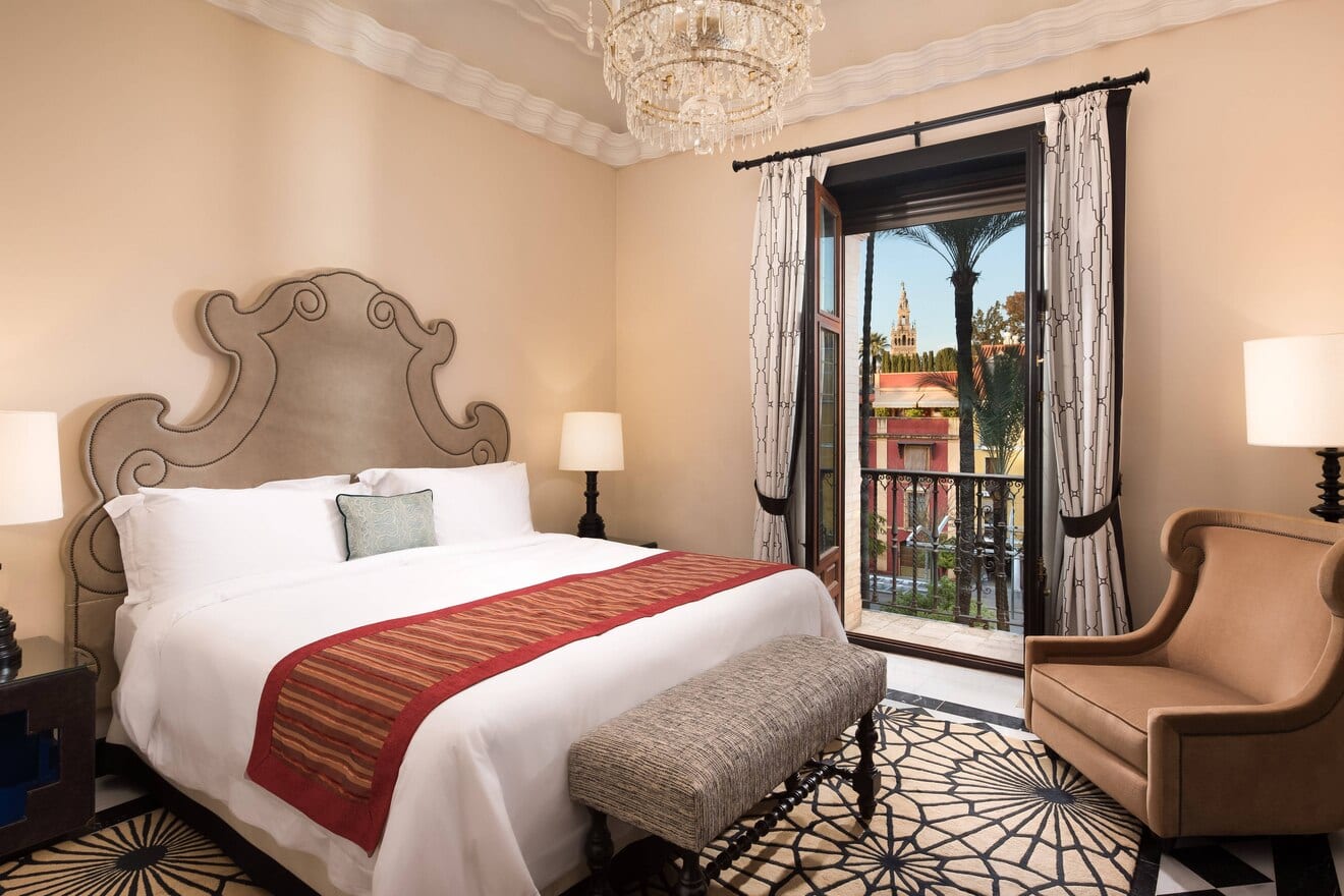 Descubra la máxima elegancia en la Suite Deluxe del Hotel Alfonso XIII de Sevilla. Disfrute de una espaciosa habitación con una cama grande, lámpara de araña y sillón, mientras admira las impresionantes vistas a una histórica torre enmarcada por frondosas palmeras desde la ventana. - ForTwo - Planes para parejas