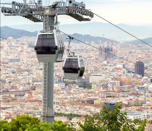 Disfrute de impresionantes vistas mientras los teleféricos sobrevuelan el paisaje urbano de Barcelona con montañas lejanas y cielos parcialmente nublados como telón de fondo. Reserve sus Entradas Teleférico de Barcelona para una aventura inigualable sobre la vibrante ciudad. - ForTwo - Planes para parejas