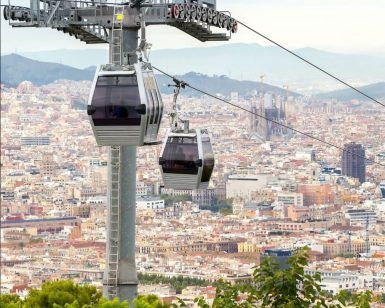 Disfrute de impresionantes vistas mientras los teleféricos sobrevuelan el paisaje urbano de Barcelona con montañas lejanas y cielos parcialmente nublados como telón de fondo. Reserve sus Entradas Teleférico de Barcelona para una aventura inigualable sobre la vibrante ciudad. - ForTwo - Planes para parejas