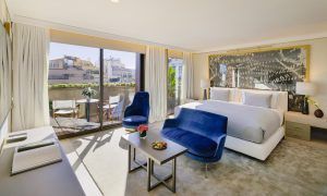 Descubra la elegancia del Hotel The One Barcelona GL, que cuenta con una espaciosa habitación con una cama grande, asientos azules y puertas de cristal que se abren a un balcón con impresionantes vistas a la ciudad. - ForTwo - Planes para parejas