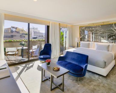 Descubra la elegancia del Hotel The One Barcelona GL, que cuenta con una espaciosa habitación con una cama grande, asientos azules y puertas de cristal que se abren a un balcón con impresionantes vistas a la ciudad. - ForTwo - Planes para parejas