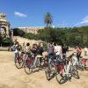 Un grupo del "Free tour por Barcelona en bicicleta" explora un parque, pasando por una gran fuente y un impresionante monumento bajo el cielo azul claro. - ForTwo - Planes para parejas