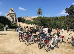 Un grupo del "Free tour por Barcelona en bicicleta" explora un parque, pasando por una gran fuente y un impresionante monumento bajo el cielo azul claro. - ForTwo - Planes para parejas