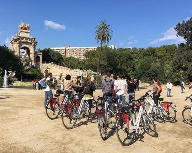 Un grupo del "Free tour por Barcelona en bicicleta" explora un parque, pasando por una gran fuente y un impresionante monumento bajo el cielo azul claro. - ForTwo - Planes para parejas
