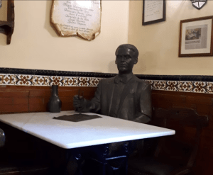 Entre azulejos decorativos y cuadros enmarcados, una estatua de bronce reposa elegantemente en una mesa de café. Ideal para reflexionar durante "Federico García Lorca: Tour guiado por Granada", que captura la esencia artística de Lorca. - ForTwo - Planes para parejas